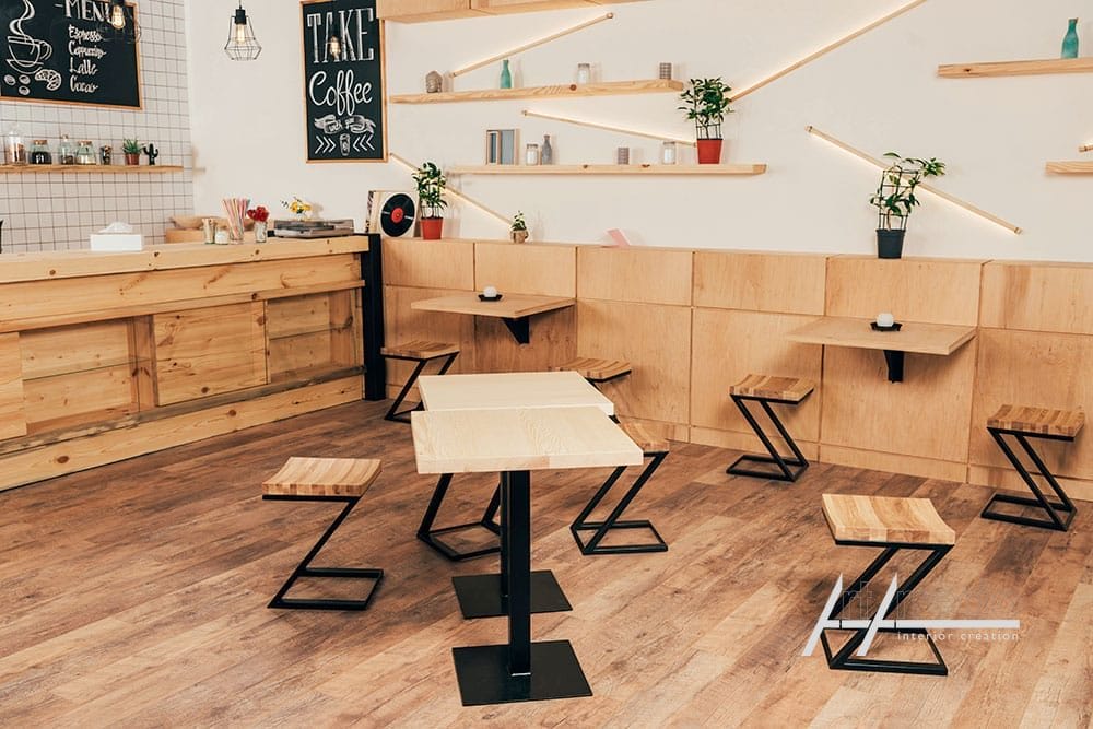 Interni moderni di una caffetteria con tavoli in legno, panche e arredamento minimalista, con scaffali con piante e menù contract su misura su lavagna.