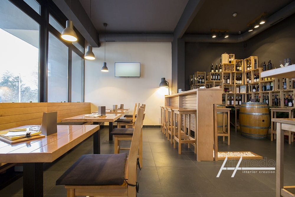 Interni moderni del ristorante con tavoli e sedie in legno su misura, portabottiglie, lampade a sospensione e grandi finestre che mostrano la luce esterna.