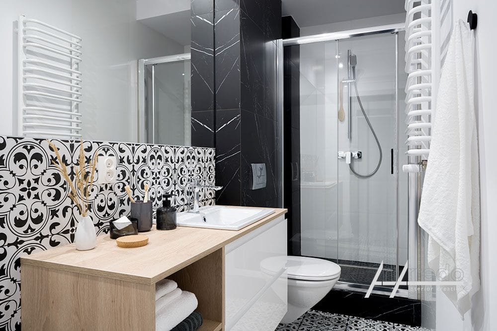 Interno del bagno moderno con piastrelle bianche e nere, doccia in vetro trasparente, WC bianco, scaldasalviette e lavabo in legno ArtArredo Piacenza.