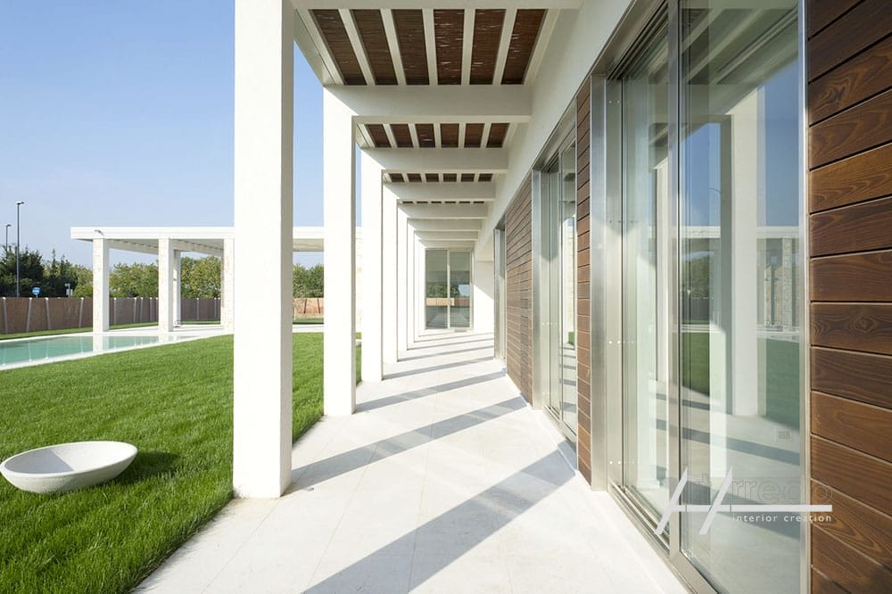 Esterno di una casa moderna con passerella a colonne, rivestimenti in legno, ampie finestre e vista su un prato verde e piscina, progettata con "arredi per privati".