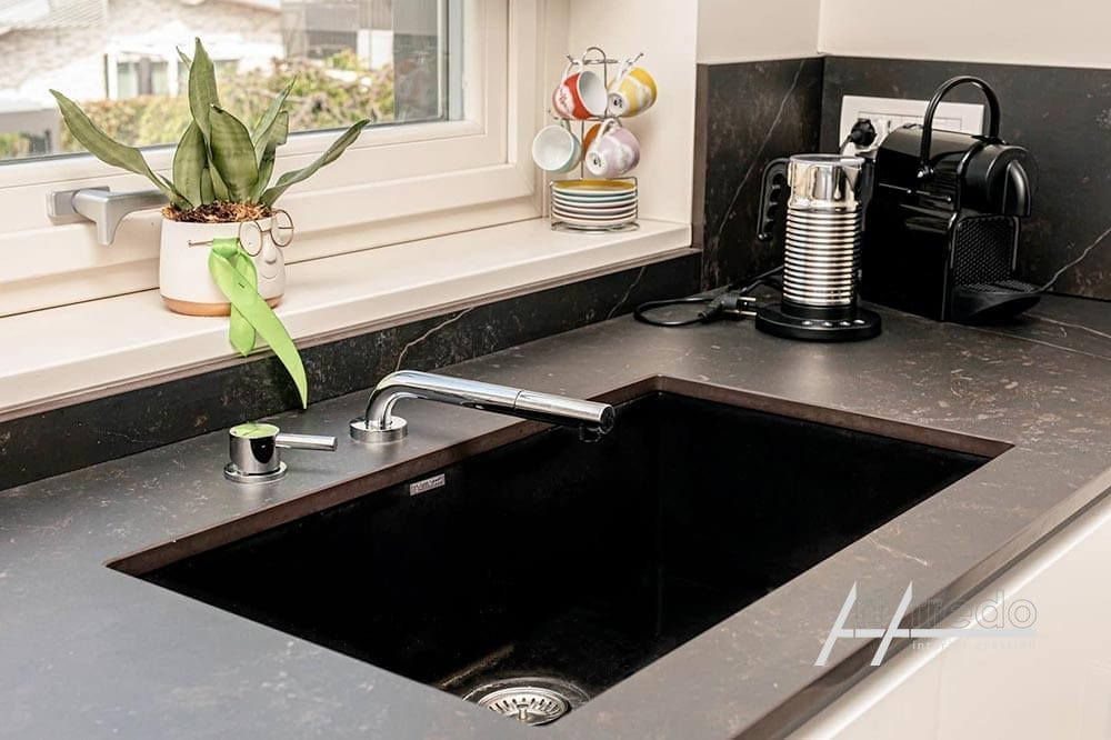 Un moderno piano di lavoro della cucina con lavello nero, macchina per il caffè e una pianta in vaso vicino a una finestra, caratterizzato da elementi di design personalizzati.