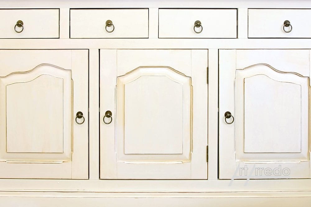 Mobile in legno bianco con ante e cassetti chiusi, caratterizzato da pomelli circolari metallici e pannelli decorativi modanati, concepito come mobili classici.