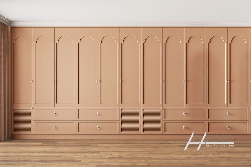 Una fila di eleganti mobili Mobili Classici in legno color pesca con dettagli ad arco, dotati di cassetti e griglie di ventilazione nella parte inferiore, appoggiati su un pavimento in legno e una parete liscia.